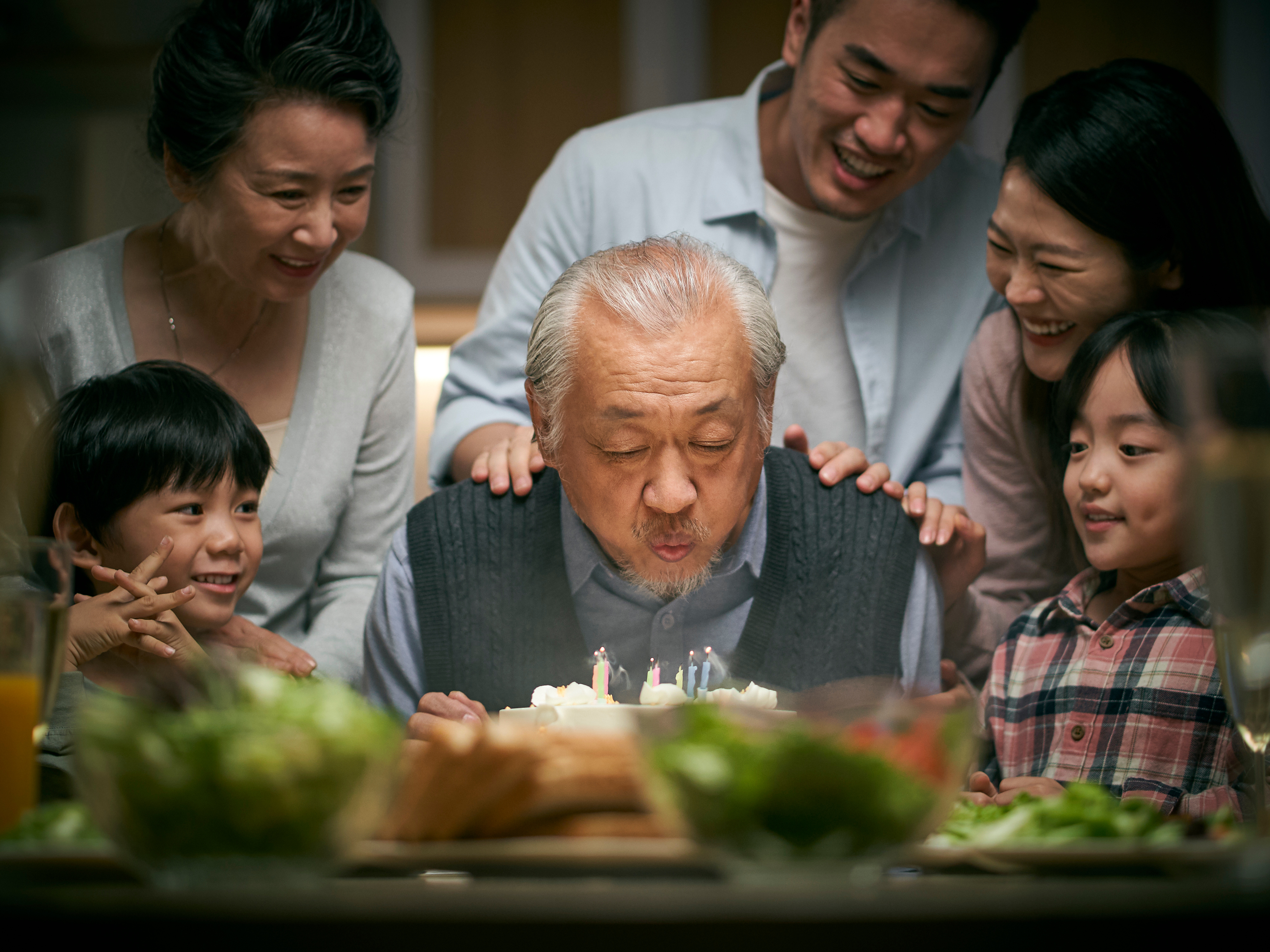 grandfather blowing out candles on a birthday cake with family around him